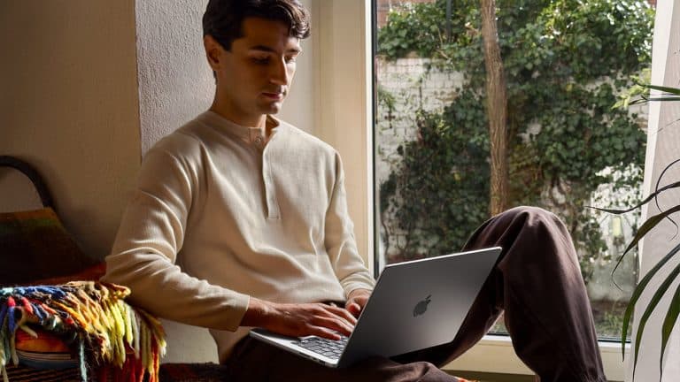 A man sits by a window with greenery outside, working on his laptop during Apple Back to School season. He wears a cream henley shirt and brown pants, relaxing on a colorful blanket in a cozy, well-lit room.