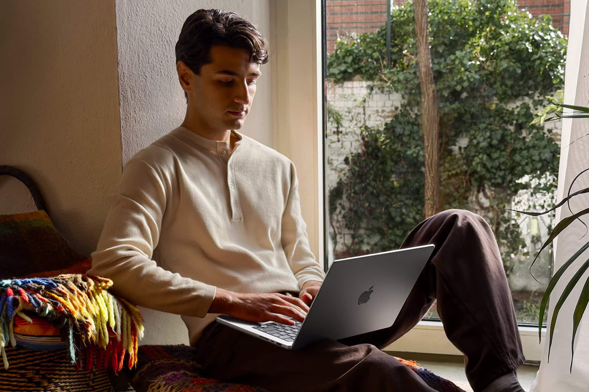 A man sits by a window with greenery outside, working on his laptop during Apple Back to School season. He wears a cream henley shirt and brown pants, relaxing on a colorful blanket in a cozy, well-lit room.
