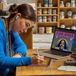 A woman wearing a blue sweater uses a stylus to draw on a tablet at a wooden desk. Behind her, a laptop from the Apple Back to School collection displays colorful digital artwork while jars of paint are visible on shelves.