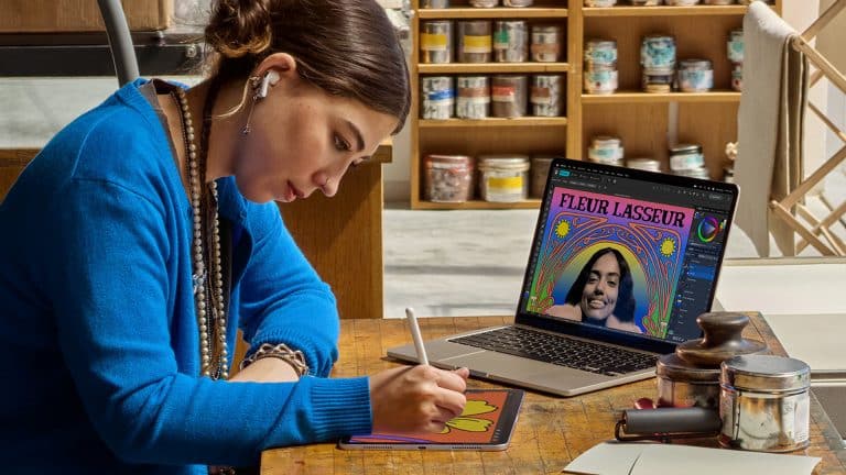 A woman wearing a blue sweater uses a stylus to draw on a tablet at a wooden desk. Behind her, a laptop from the Apple Back to School collection displays colorful digital artwork while jars of paint are visible on shelves.
