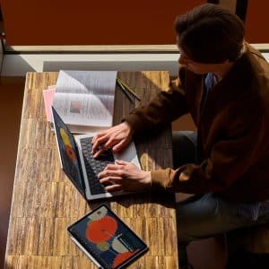 A person sits at a wooden table using a laptop with data charts on the screen, preparing for Apple Back to School. An open book, a pencil, and a tablet displaying graphs are also on the table as sunlight streams in through nearby windows.