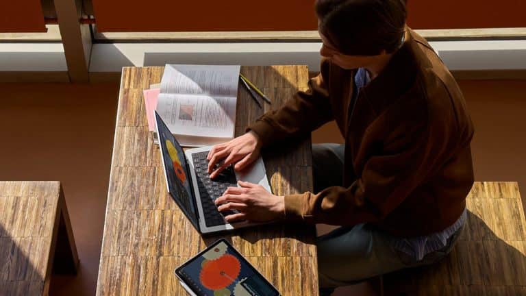 A person sits at a wooden table using a laptop with data charts on the screen, preparing for Apple Back to School. An open book, a pencil, and a tablet displaying graphs are also on the table as sunlight streams in through nearby windows.