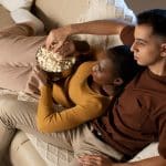 A couple sits close together on a beige couch, sharing a bowl of popcorn during an Apple TV weekend. The woman in a mustard sweater leans on the man in a brown shirt as they look ahead, appearing relaxed and comfortable.