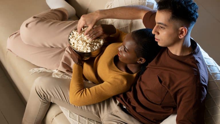 A couple sits close together on a beige couch, sharing a bowl of popcorn during an Apple TV weekend. The woman in a mustard sweater leans on the man in a brown shirt as they look ahead, appearing relaxed and comfortable.