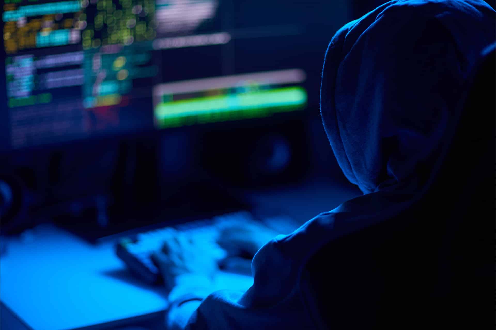 A person in a hooded sweatshirt types on a keyboard in a dark room, illuminated by blue light from multiple computer screens displaying code and data related to the Foxconn cyberattack.