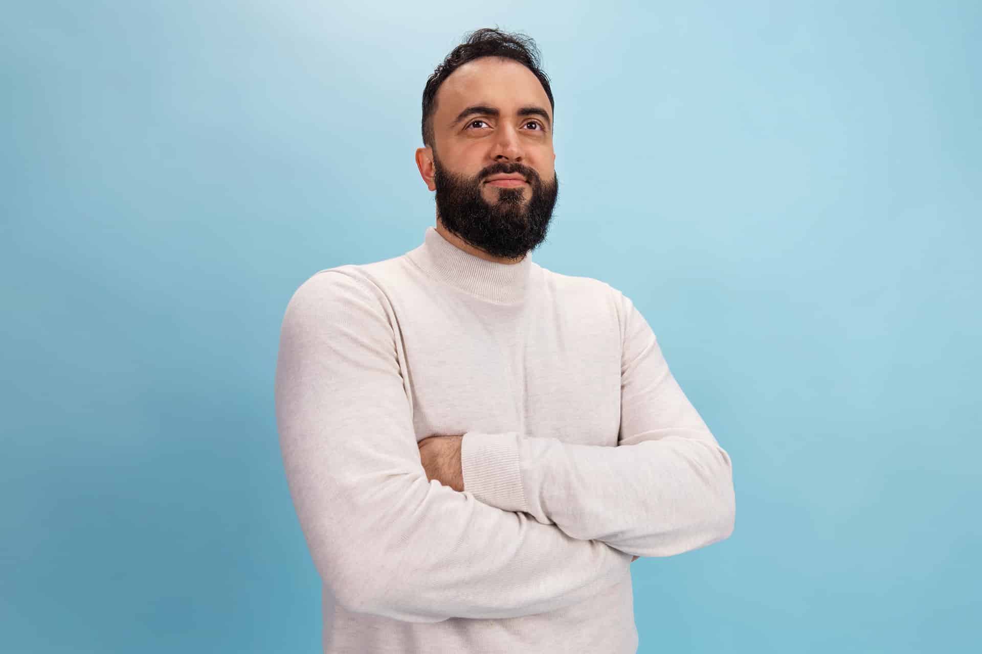 A man with a beard and dark hair stands confidently with arms crossed, wearing a light-colored turtleneck sweater—reflecting the innovation of Detroit developers and the spirit of the Apple Developer Academy—against a plain light blue background.