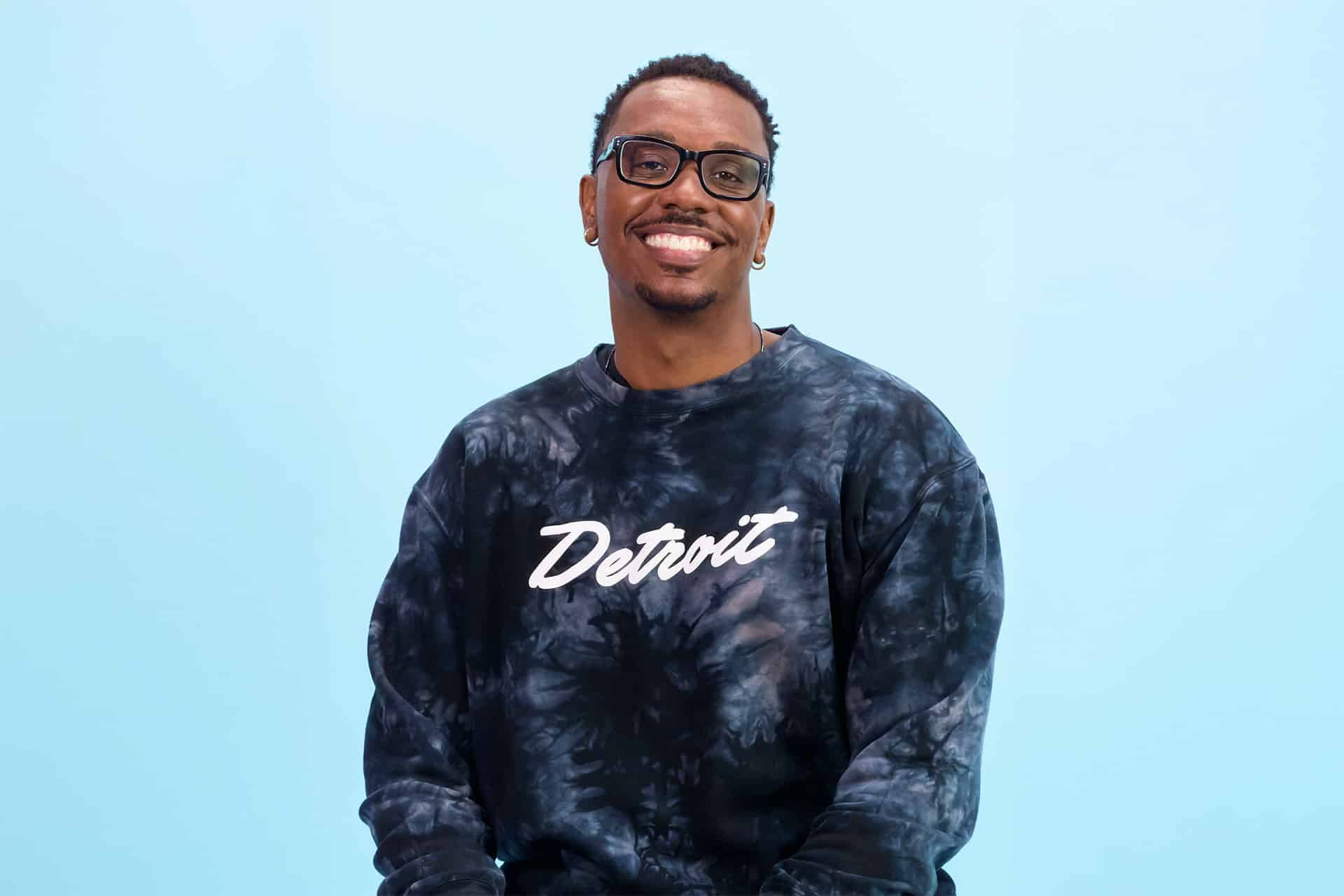 A smiling man wearing glasses and a black tie-dye "Detroit" sweatshirt, sitting against a plain light blue background—an advocate for Apple education and app development inspired by his Michigan State University experience.