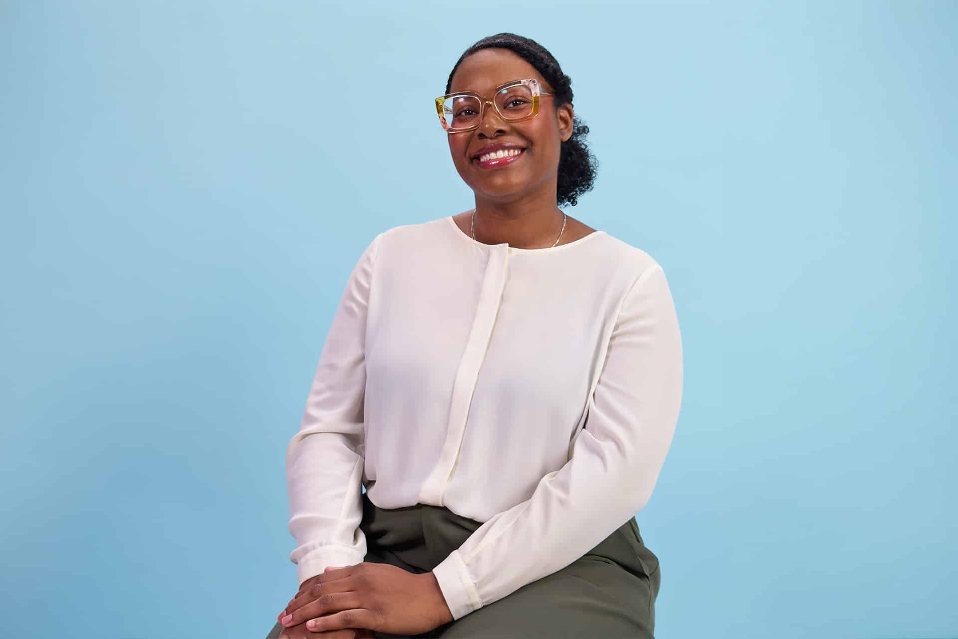 A woman wearing glasses, a white blouse, and dark pants sits and smiles against a plain light blue background, representing the spirit of Detroit developers and their passion for app development.