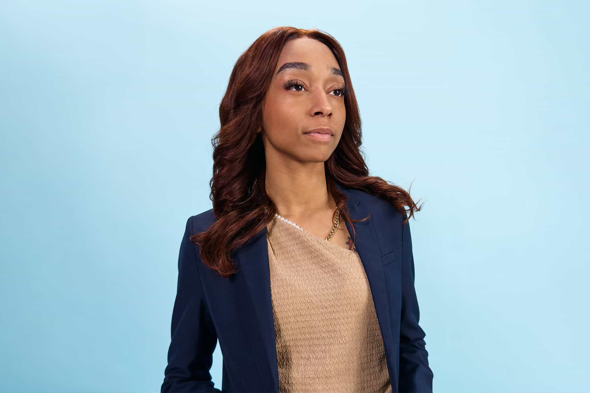 A woman with long brown hair wearing a navy blazer and beige top stands against a light blue background, looking confidently into the distance—an aspiring innovator from the Apple Developer Academy at Michigan State University.
