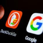 A close-up of a smartphone screen shows the DuckDuckGo iPhone app icon, featuring a duck, next to the Google app icon with its colorful "G" logo. A finger is partially visible near the DuckDuckGo icon.