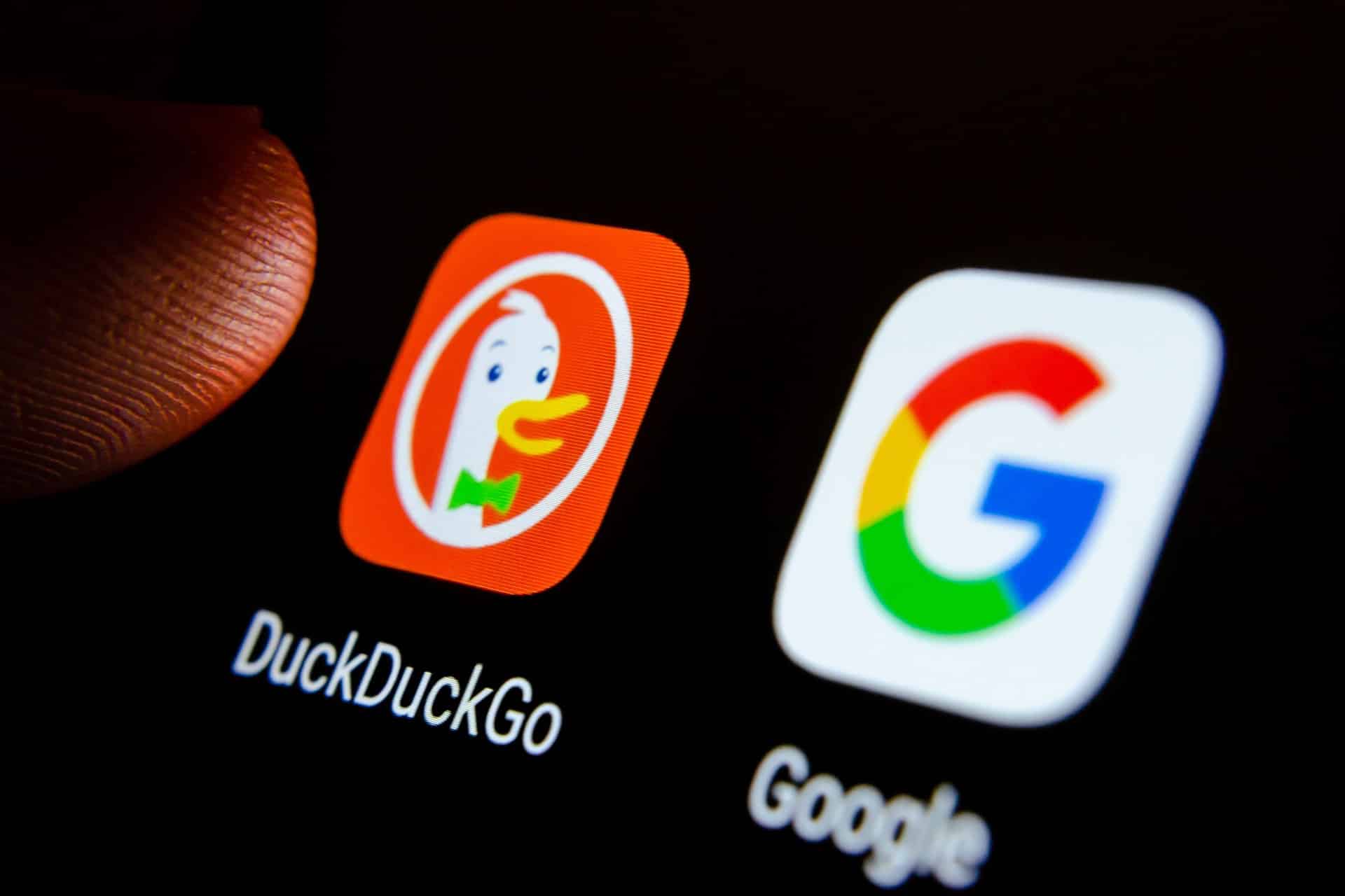 A close-up of a smartphone screen shows the DuckDuckGo iPhone app icon, featuring a duck, next to the Google app icon with its colorful "G" logo. A finger is partially visible near the DuckDuckGo icon.