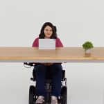 A woman in a wheelchair sits at a wooden desk with a tablet, a small potted plant, and an iPhone with Eye Tracking features, facing forward against a plain white background.