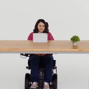 A woman in a wheelchair sits at a wooden desk with a tablet, a small potted plant, and an iPhone with Eye Tracking features, facing forward against a plain white background.
