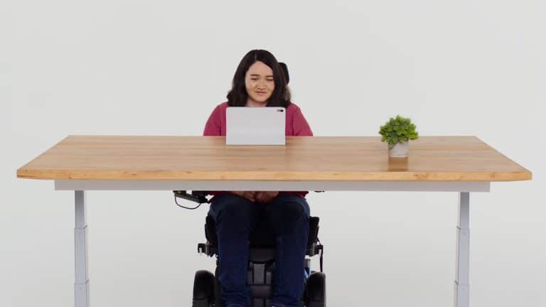 A woman in a wheelchair sits at a wooden desk with a tablet, a small potted plant, and an iPhone with Eye Tracking features, facing forward against a plain white background.