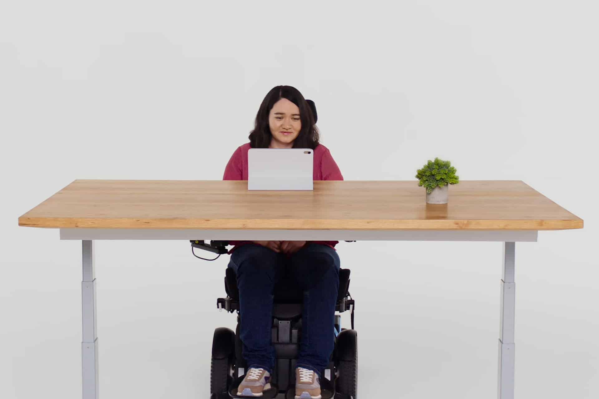 A woman in a wheelchair sits at a wooden desk with a tablet, a small potted plant, and an iPhone with Eye Tracking features, facing forward against a plain white background.