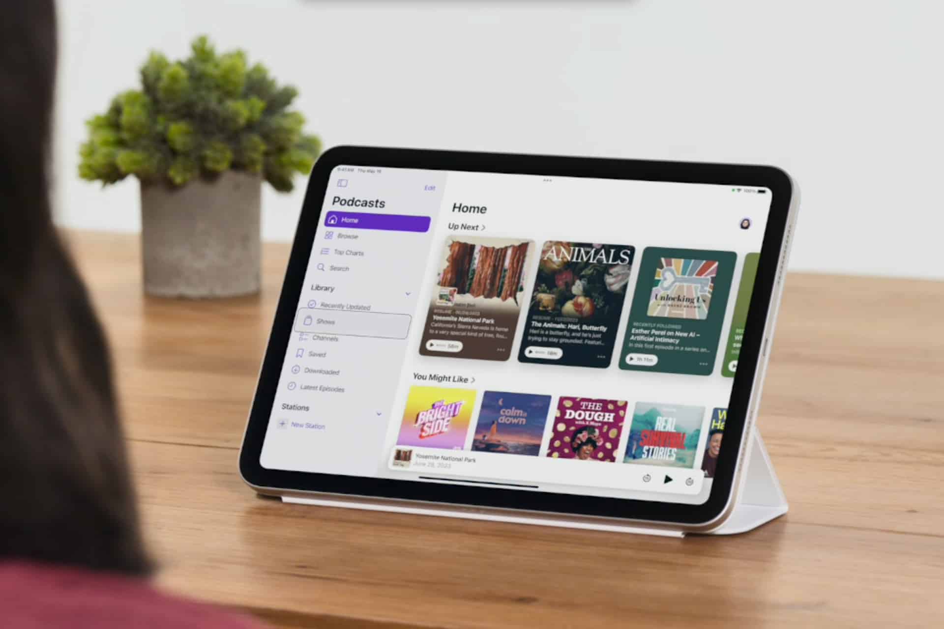A person sits at a table viewing a tablet displaying a podcast app with colorful covers and categories visible, while iPhone Eye Tracking technology could further enhance their browsing experience. A small potted plant is in the background.