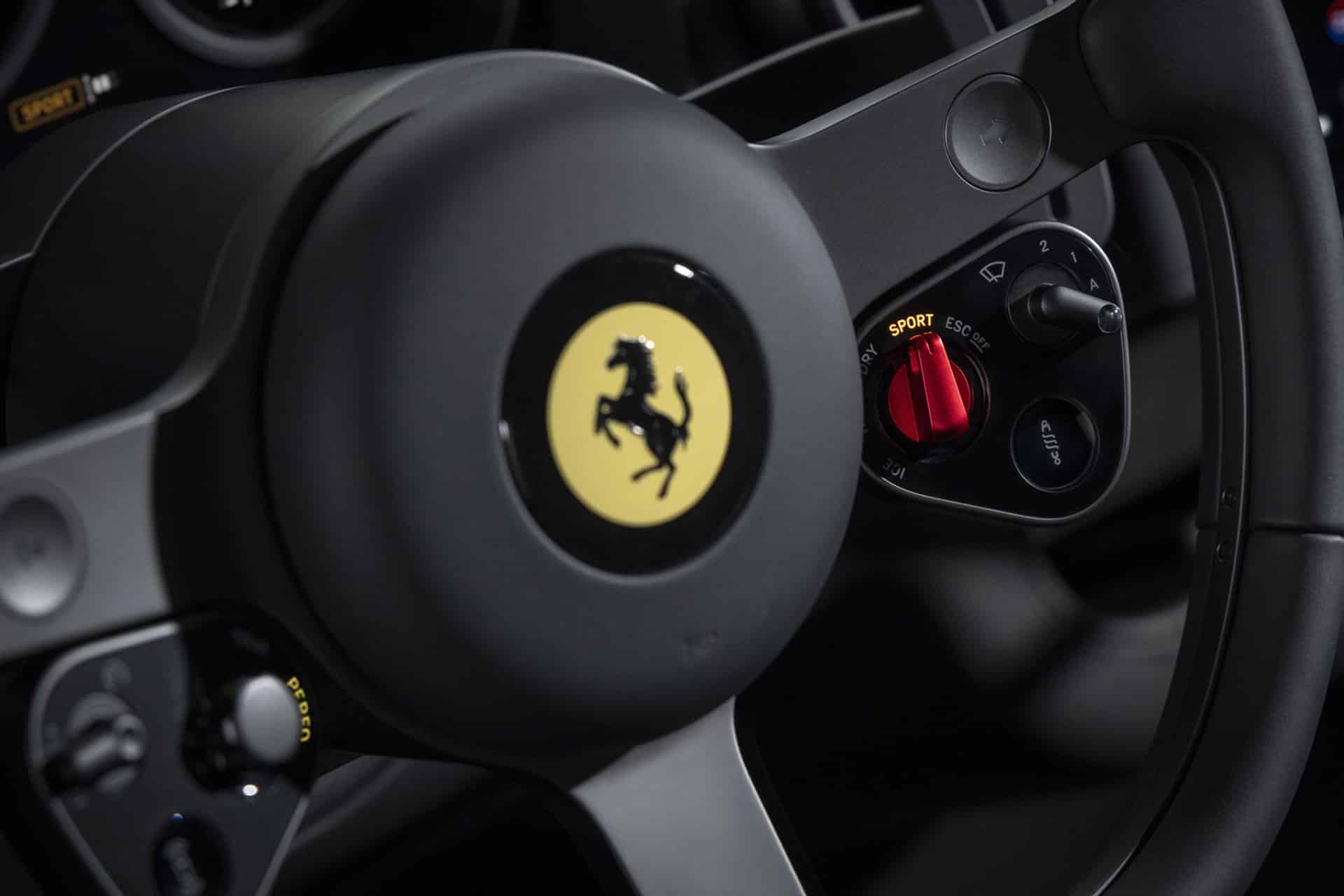 Close-up of a Ferrari steering wheel, featuring the iconic prancing horse logo and a red driving mode selector in Sport mode, with Ferrari Luce CarPlay seamlessly integrated into the modern design.