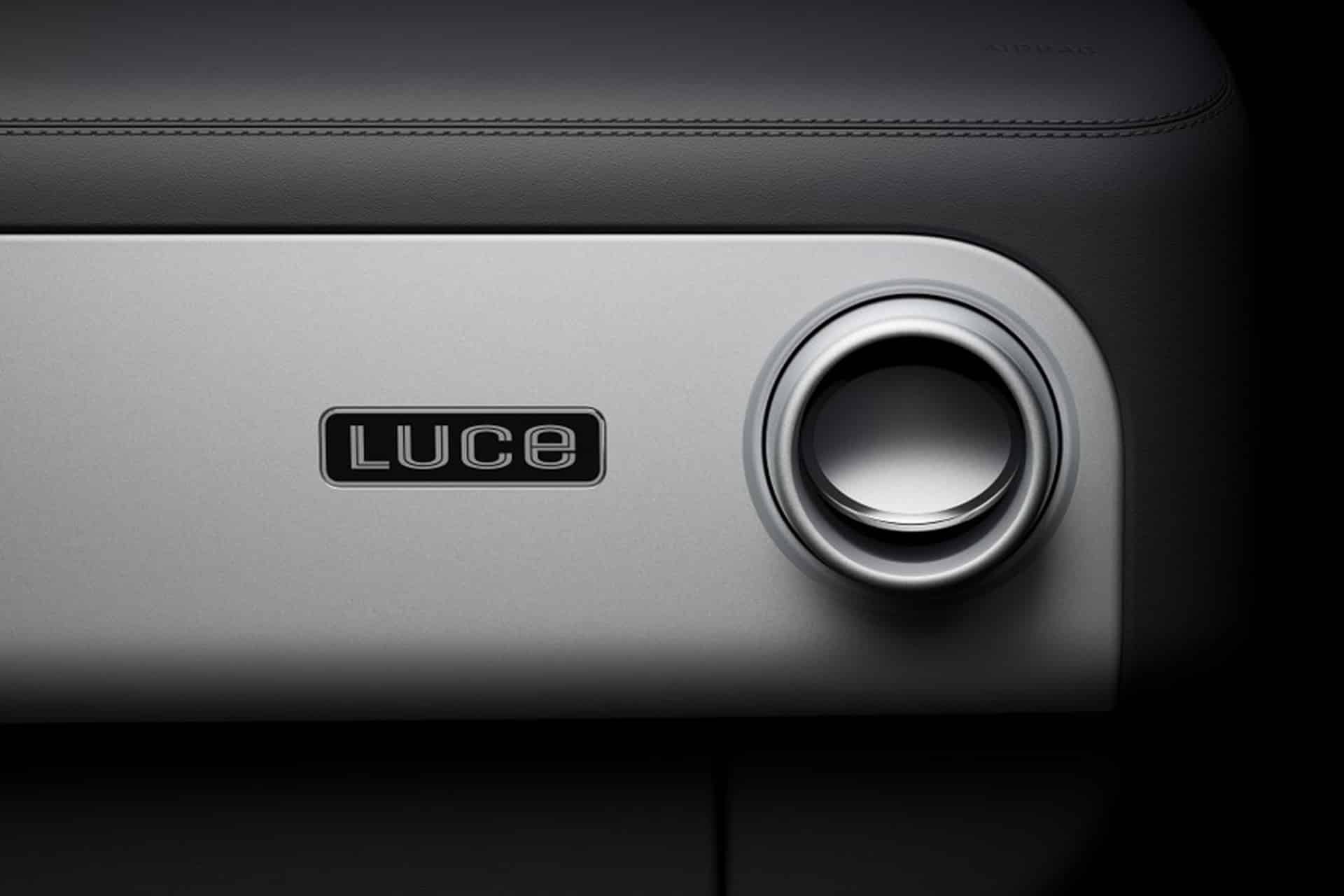 Close-up of a car dashboard featuring a round air vent and a "LUCE" badge on a smooth, light panel, showcasing Ferrari Luce CarPlay integration. The surface above is dark and textured, evoking the minimalist style of Jony Ive.