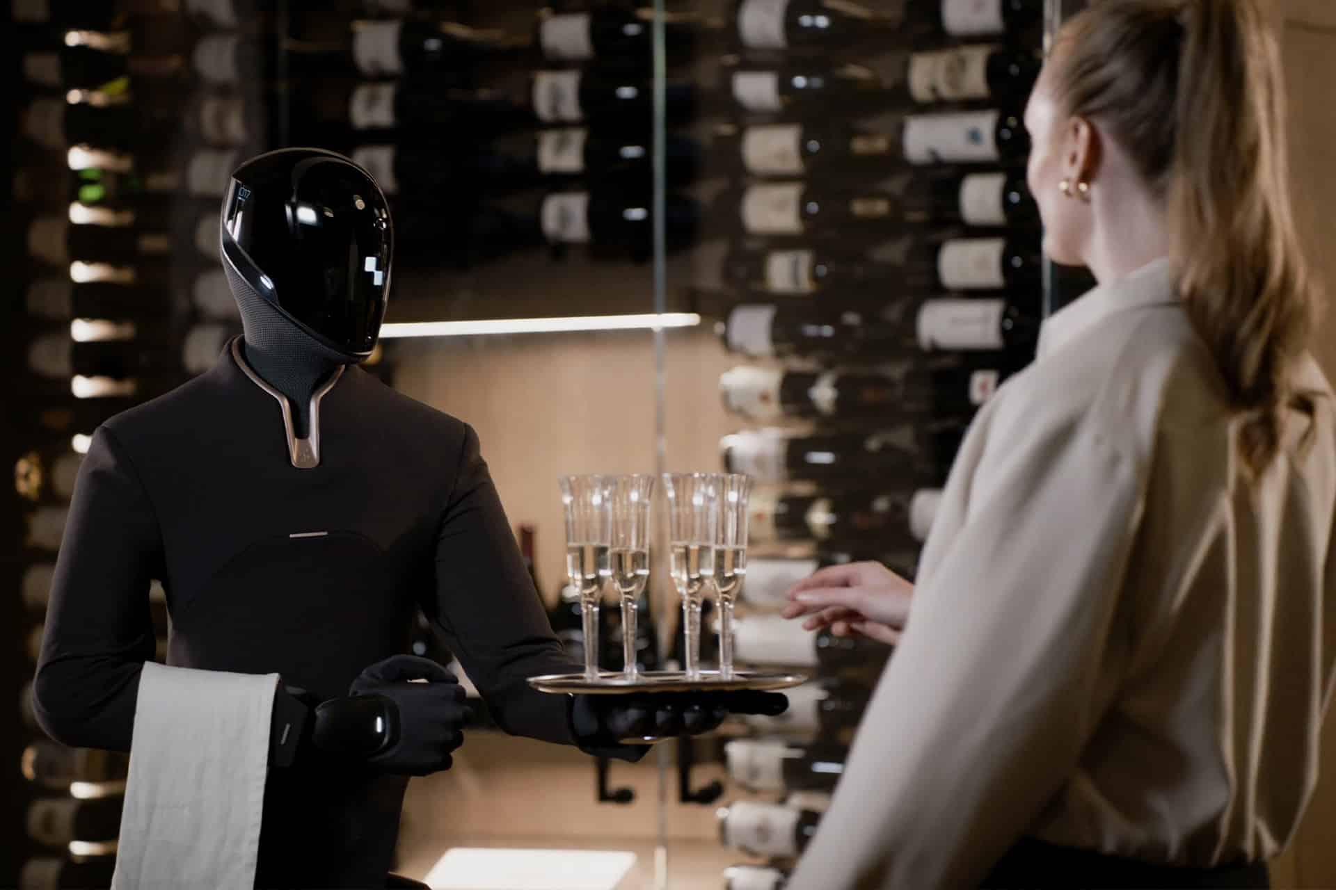 A humanoid robot by Figure AI in a sleek black uniform serves champagne glasses on a tray to a woman in a wine cellar lined with bottles. The woman reaches for a glass as the F.03 model holds a towel, resembling an attentive waiter.