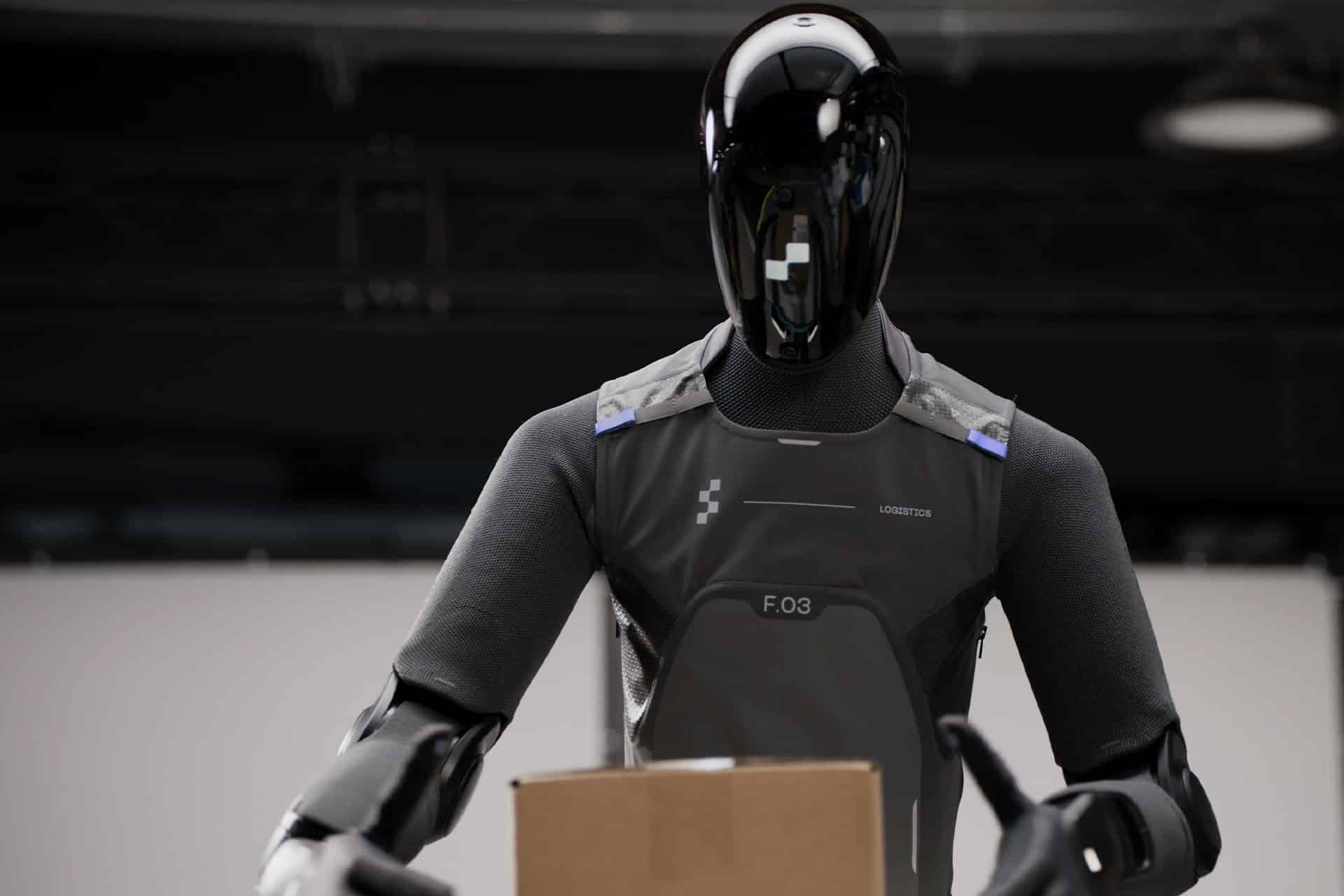 A sleek, black Figure AI humanoid robot wearing a gray suit stands indoors, holding a cardboard box. The robot, built with advanced physical AI and segmented arms, has a shiny, featureless face. The industrial background is blurred.