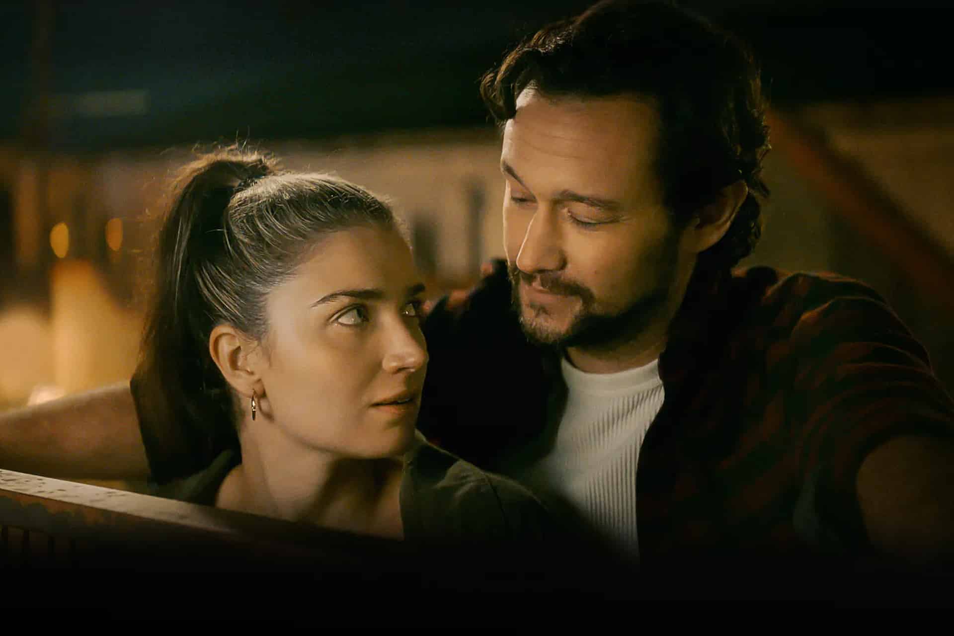 A woman with long brown hair in a ponytail looks up at a man with wavy hair and a beard as he gazes at her, both seated closely together in a warmly lit setting, capturing an intimate moment reminiscent of Apple TV Movies.