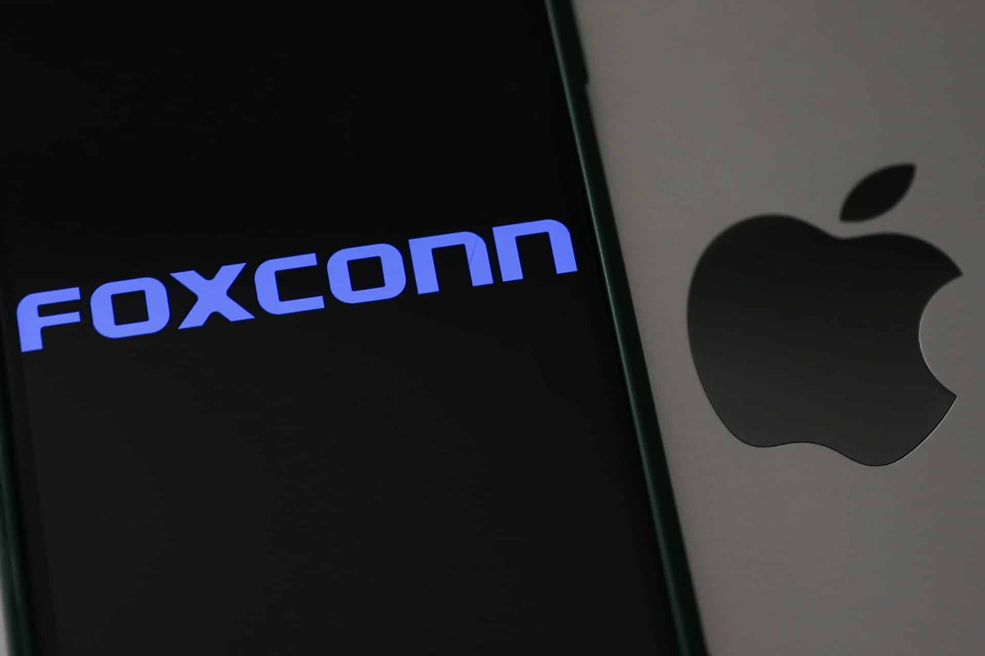 A smartphone screen displays the blue Foxconn logo next to a close-up of the Apple logo on a silver device, referencing concerns about the recent Foxconn cyberattack.