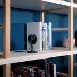 A black indoor security camera with Home Activity Zones sits on a white shelf among books, a gray smart speaker, and a pink cup, against a blue wall.