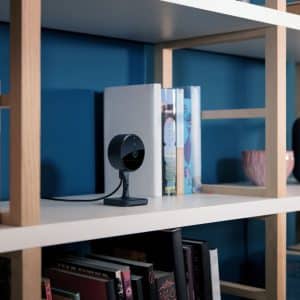 A black indoor security camera with Home Activity Zones sits on a white shelf among books, a gray smart speaker, and a pink cup, against a blue wall.