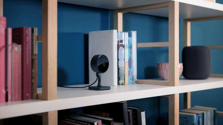 A black indoor security camera with Home Activity Zones sits on a white shelf among books, a gray smart speaker, and a pink cup, against a blue wall.