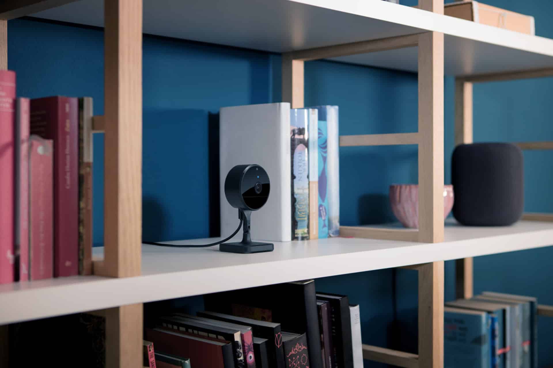 A black indoor security camera with Home Activity Zones sits on a white shelf among books, a gray smart speaker, and a pink cup, against a blue wall.