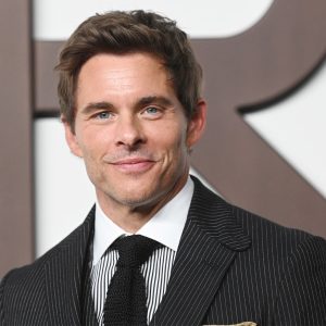 A man with short brown hair in a dark pinstripe suit, patterned tie, and pocket square is smiling at the camera, standing in front of a white background with large, dark, blurred letters—a look reminiscent of Apple TV's Disavowed.