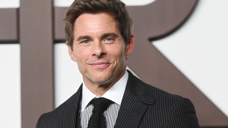 A man with short brown hair in a dark pinstripe suit, patterned tie, and pocket square is smiling at the camera, standing in front of a white background with large, dark, blurred letters—a look reminiscent of Apple TV's Disavowed.