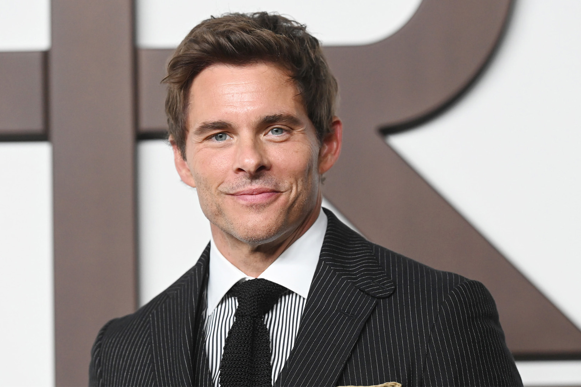 A man with short brown hair in a dark pinstripe suit, patterned tie, and pocket square is smiling at the camera, standing in front of a white background with large, dark, blurred letters—a look reminiscent of Apple TV's Disavowed.