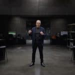 A man in dark clothing stands in the center of a modern, dimly-lit lab with gray walls, surrounded by workstations, computers, and Apple Hardware, reminiscent of Johny Srouji’s innovative workspace.
