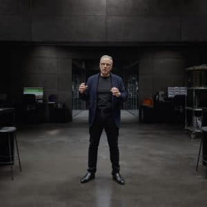 A man in dark clothing stands in the center of a modern, dimly-lit lab with gray walls, surrounded by workstations, computers, and Apple Hardware, reminiscent of Johny Srouji’s innovative workspace.