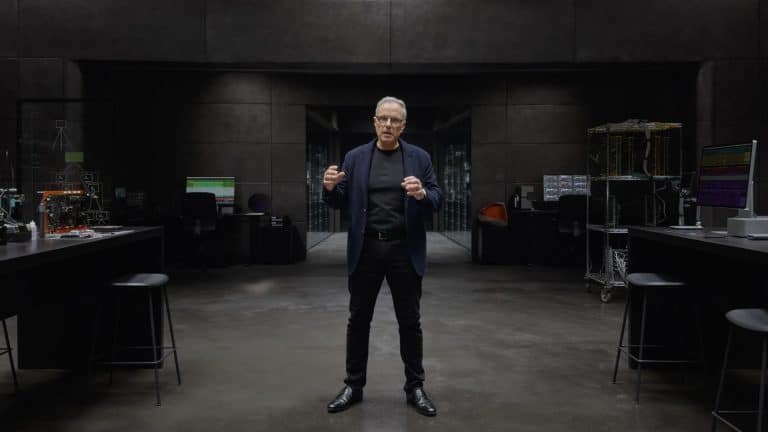 A man in dark clothing stands in the center of a modern, dimly-lit lab with gray walls, surrounded by workstations, computers, and Apple Hardware, reminiscent of Johny Srouji’s innovative workspace.
