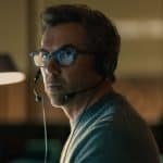 A man wearing glasses and a headset sits in a dimly lit room, looking intently to the side. He appears focused, with a soft light illuminating his face—reminiscent of scenes from Last Seen Apple TV. Blurred background elements are visible.