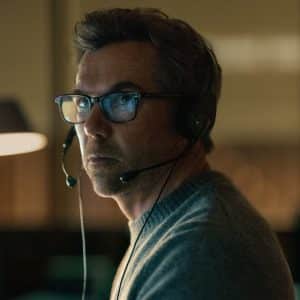 A man wearing glasses and a headset sits in a dimly lit room, looking intently to the side. He appears focused, with a soft light illuminating his face—reminiscent of scenes from Last Seen Apple TV. Blurred background elements are visible.