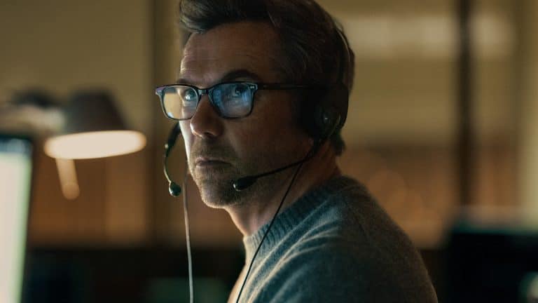 A man wearing glasses and a headset sits in a dimly lit room, looking intently to the side. He appears focused, with a soft light illuminating his face—reminiscent of scenes from Last Seen Apple TV. Blurred background elements are visible.