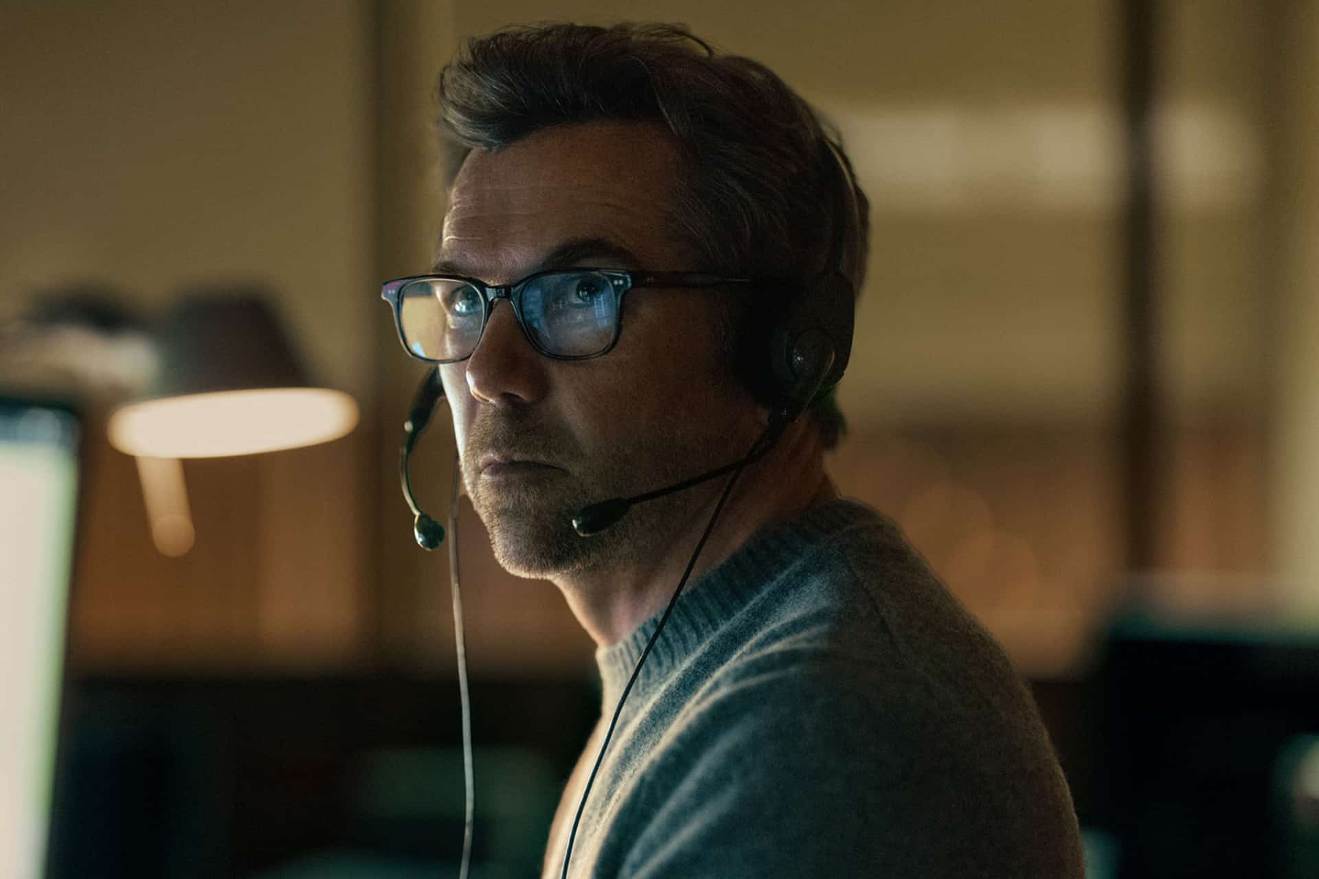 A man wearing glasses and a headset sits in a dimly lit room, looking intently to the side. He appears focused, with a soft light illuminating his face—reminiscent of scenes from Last Seen Apple TV. Blurred background elements are visible.