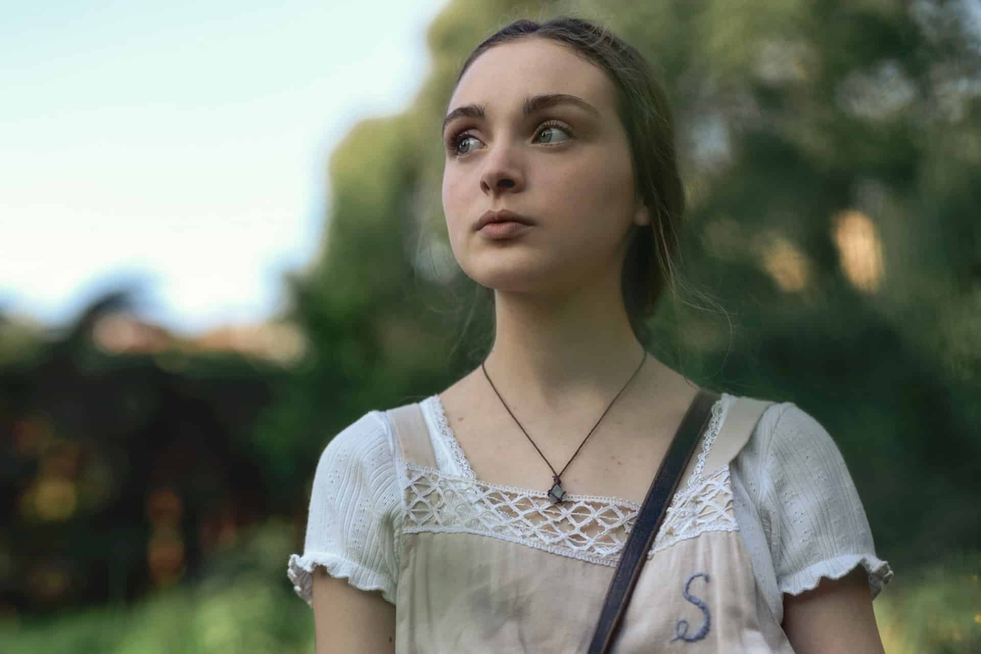 A young woman with light brown hair tied back, wearing a white blouse and a cream-colored dress with lace details, stands outdoors with a thoughtful expression—she looks as if she could be a character from Last Seen Apple TV. Green foliage and soft sunlight blur behind her.