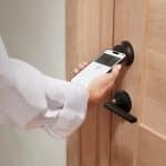 A person is holding a smartphone near a HomeKit smart lock, appearing to unlock the wooden door using a digital key or mobile access app.
