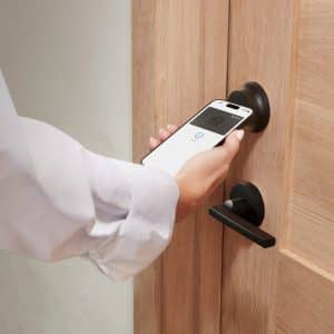 A person is holding a smartphone near a HomeKit smart lock, appearing to unlock the wooden door using a digital key or mobile access app.