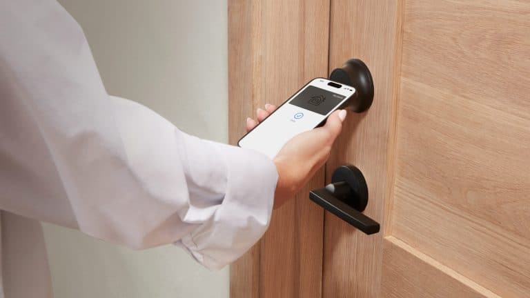 A person is holding a smartphone near a HomeKit smart lock, appearing to unlock the wooden door using a digital key or mobile access app.