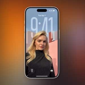 A smartphone running iOS 26 Adaptive Time displays its lock screen, showing the time 9:41 and date Tuesday, April 1. The wallpaper features a blonde woman in a black outfit posing outdoors against a pink wall and blue sky.