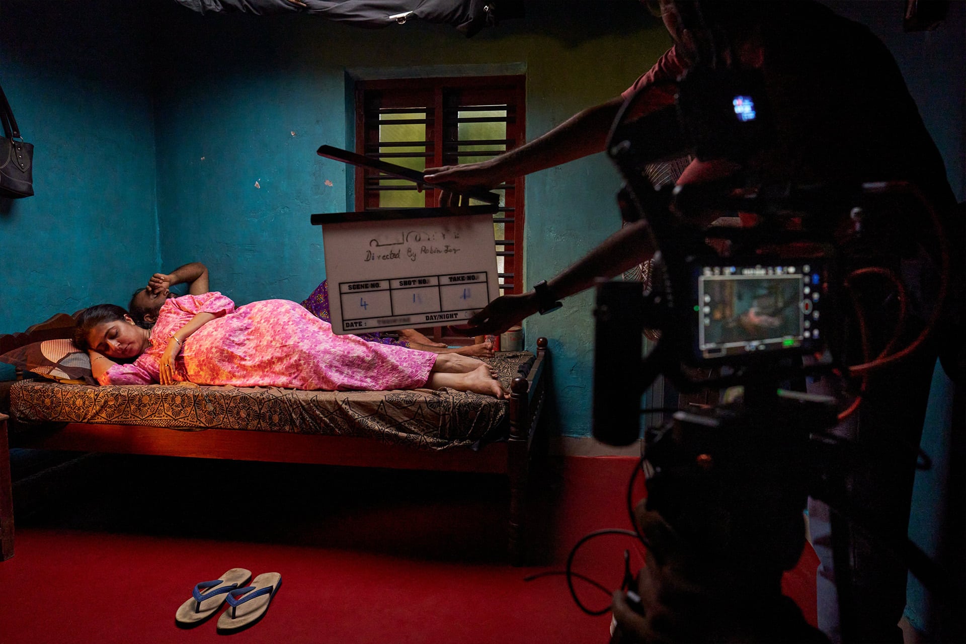 A woman in a pink dress lies on a bed in a dimly lit room, as a film crew prepares to shoot a scene. A clapperboard is held in front of her; filmed on iPhone, with a camera and monitor visible in the foreground.