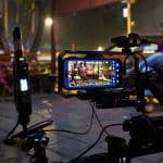 A smartphone mounted on filming equipment records two women sitting on wooden benches outdoors at night, with one woman in focus on the phone screen and the other blurred in the background—filmed on iPhone.