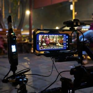 A smartphone mounted on filming equipment records two women sitting on wooden benches outdoors at night, with one woman in focus on the phone screen and the other blurred in the background—filmed on iPhone.