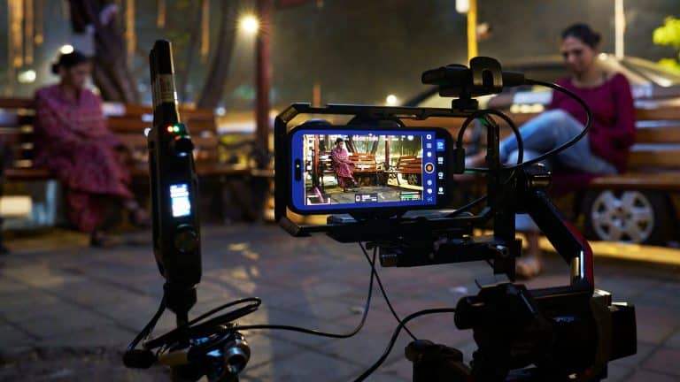 A smartphone mounted on filming equipment records two women sitting on wooden benches outdoors at night, with one woman in focus on the phone screen and the other blurred in the background—filmed on iPhone.