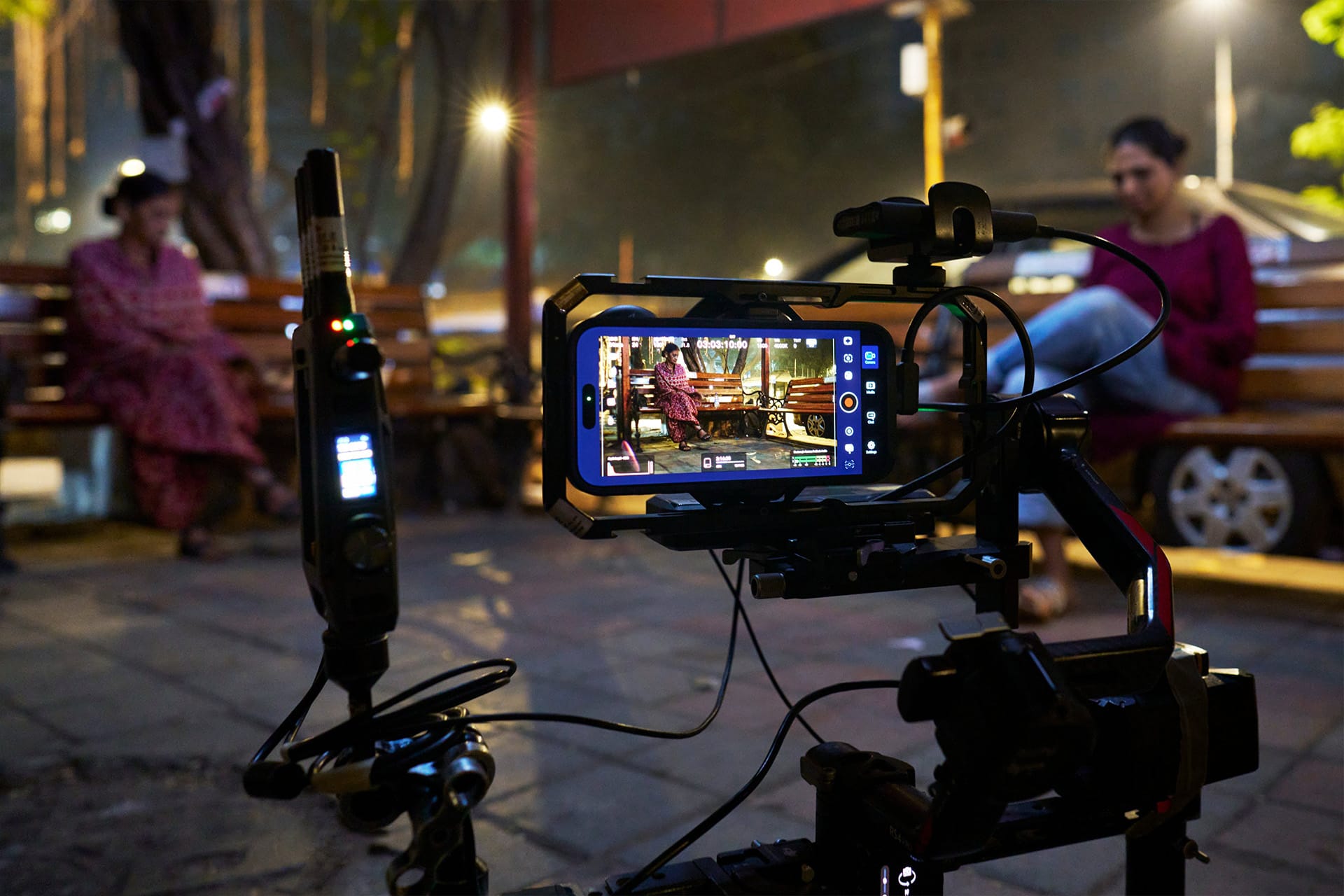 A smartphone mounted on filming equipment records two women sitting on wooden benches outdoors at night, with one woman in focus on the phone screen and the other blurred in the background—filmed on iPhone.